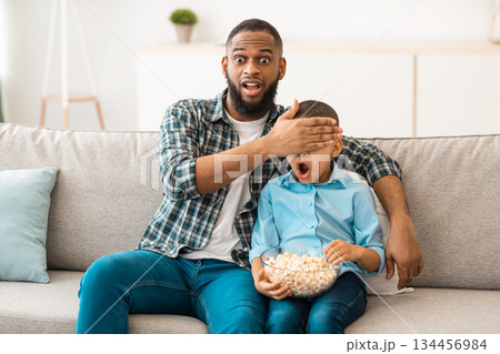 Parental Control. Shocked African Dad And Son Watching TV Together, Father Covering Kid's Eyes Looking At Questionable Content Sitting On Couch At Home. Children Tv Programming Problem Concept 134456984