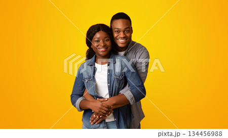 Portrait of happy african american lovers hugging and smiling at camera while standing together isolated over yellow background, romantic black guy cuddling his girlfriend from back, copy space 134456988