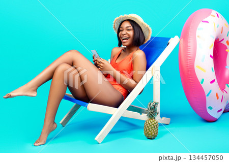 Lovely black woman relaxing in lounge chair, using smartphone, chatting to friend online on beach, blue background. Cheerful young lady communicating on web during summer vacation Lovely black woman relaxing in lounge chair, using smartphone, chatting to friend online on beach, blue background. Cheerful young lady communicating on web during summer vacation 134457050