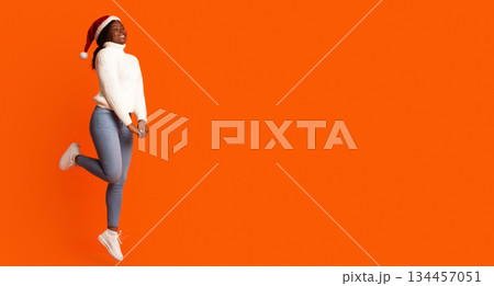 Christmas Mood. Portrait Of Happy Jumping Black Lady In Santa Hat Over Orange Background, Cheerful African American Woman Having Fun, Enjoying Xmas And New Year Holidays, Full Length Shot, Copy Space Christmas Mood. Portrait Of Happy Jumping Black Lady In Santa Hat Over Orange Background, Cheerful African American Woman Having Fun, Enjoying Xmas And New Year Holidays, Full Length Shot, Copy Space 134457051
