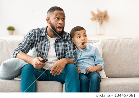 Television Programming Problem. Shocked African Father And Son Watching Questionable Content On TV Together Sitting On Couch With Opened Mouth At Home. Parental Control Concept 134457061