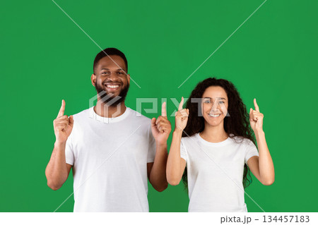 Diverse Couple Pointing Fingers Up Posing Standing In Studio Over Green Background, Smiling To Camera. Multiethnic Man And Woman Showing Something Above Their Heads. Look Upward, Advertisement Banner 134457183