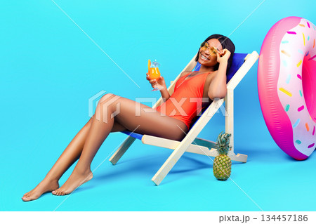 Full length portrait of young black woman chilling in lounge chair with yummy tropical cocktail on blue studio background. Relaxed millennial lady in swimwear enjoying her summer vacation Full length portrait of young black woman chilling in lounge chair with yummy tropical cocktail on blue studio background. Relaxed millennial lady in swimwear enjoying her summer vacation 134457186