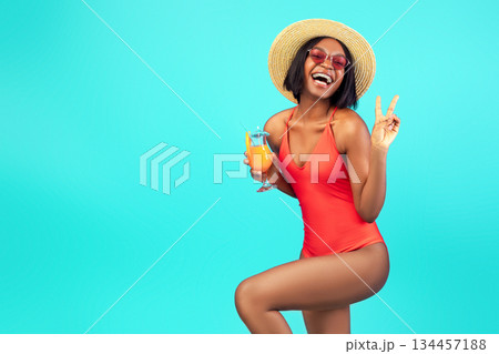 Happy black woman in swimsuit enjoying exotic summer cocktail and showing peace sign at camera, posing over blue studio background. Playful African American lady having fun on beach vacation Happy black woman in swimsuit enjoying exotic summer cocktail and showing peace sign at camera, posing over blue studio background. Playful African American lady having fun on beach vacation 134457188