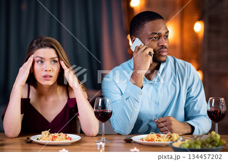 Woman looking unhappy on date while her boyfriend talking on cellphone in restaurant, paying no attention to her and busy using his mobile phone. Multiracial couple dining together at Valentine's day 134457202