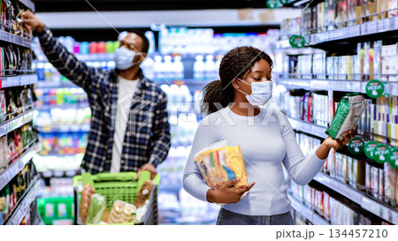 Young black woman and man in face masks choosing food products at supermarket during covid quarantine. Millennial couple wearing protection while shopping for groceries at mall Young black woman and man in face masks choosing food products at supermarket during covid quarantine. Millennial couple wearing protection while shopping for groceries at mall 134457210