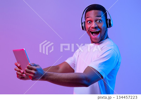 Portrait Of Overjoyed Black Guy in Wireless Headphones With Digital Tablet, Cheerful African Man Using Tab Computer And Looking At Camera With Excitement, Standing In Neon Light Over Purple Background Portrait Of Overjoyed Black Guy in Wireless Headphones With Digital Tablet, Cheerful African Man Using Tab Computer And Looking At Camera With Excitement, Standing In Neon Light Over Purple Background 134457223