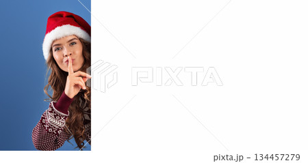 Christmas promotion. Attractive young woman in Santa hat peeking out from behind empty banner and making SILENCE gesture, blue studio background. Space for your design. Panorama Christmas promotion. Attractive young woman in Santa hat peeking out from behind empty banner and making SILENCE gesture, blue studio background. Space for your design. Panorama 134457279
