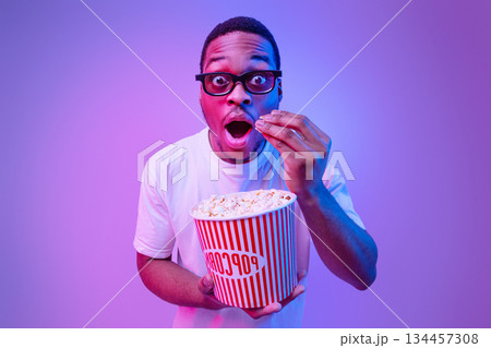Surprised African Man In 3d Glasses Holding Bucket With Popcorn And Enjoying Movie In Cinema, Standing Under Neon Lighting, Funny Black Guy Looking At Camera With Amazement, Studio Background 134457308