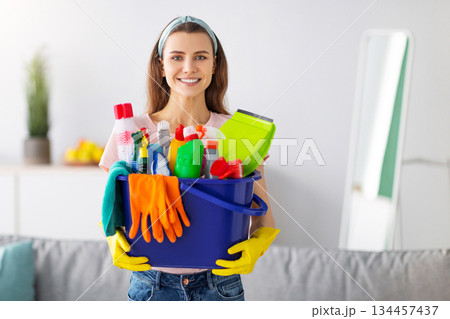 Joyful young maid holding bucket full of cleaning supplies at living room. Happy young housewife with detergents looking at camera and smiling, performing household chores with enjoyment Joyful young maid holding bucket full of cleaning supplies at living room. Happy young housewife with detergents looking at camera and smiling, performing household chores with enjoyment 134457437
