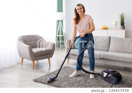 Happy young housemaid tidying her flat, vacuuming rug, indoors. Cheerful millennial lady doing household chores, hoovering carpet. Professional cleaning service concept 134457439