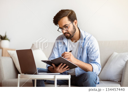 Young Arab Freelancer Man Taking Notes To Notepad And Working On Laptop At Home Office, Millennial Eastern Guy In Eyeglasses Planning Business Schedule, Sitting On Couch In Living Room, Free Space 134457440