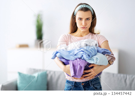 Unhappy millennial housewife holding clothes for washing or ironing, unwilling to do domestic duties, indoors. Portrait of young woman sick and tired of laundry, looking at camera and sulking 134457442