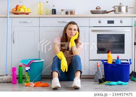 Exhausted young housewife sitting on floor at kitchen, surrounded by cleaning supplies, tired from housework. Overworked woman needing break from domestic duties, cannot stand doing house chores Exhausted young housewife sitting on floor at kitchen, surrounded by cleaning supplies, tired from housework. Overworked woman needing break from domestic duties, cannot stand doing house chores 134457475