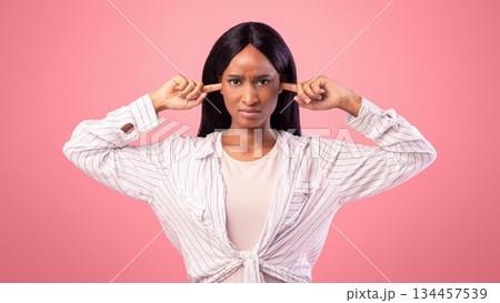 Upset black lady closing her ears with fingers, protecting from loud noise on pink studio background. African American woman not willing to listen, ignoring someone, feeling stressed and unhappy 134457539