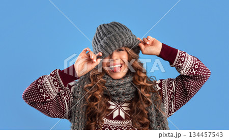 Silly young woman in warm winter clothes pulling down knitted beanie hat and sticking out her tongue on blue studio background, panorama. Lovely lady in woolen outfit having fun 134457543