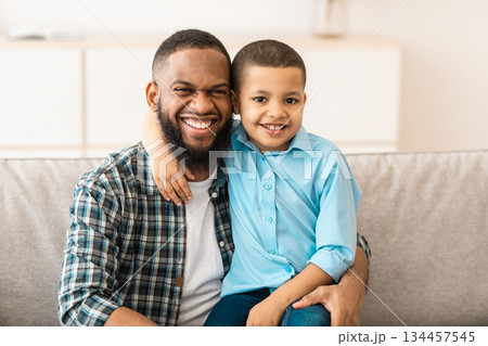 Father's Day. Happy African American Daddy And His Little Son Hugging Posing Together Embracing And Smiling To Camera Sitting On Couch At Home. Portrait Of Happy Dad And Kid Boy. Fatherhood Concept Father's Day. Happy African American Daddy And His Little Son Hugging Posing Together Embracing And Smiling To Camera Sitting On Couch At Home. Portrait Of Happy Dad And Kid Boy. Fatherhood Concept 134457545