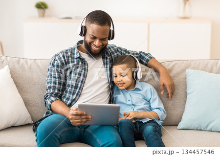 African American Father And Son Using Digital Tablet Wearing Wireless Headphones Watching Cartoons Online Together Sitting On Couch At Home. Family Weekend Leisure, Gadgets And Modern Lifestyle 134457546