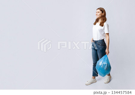 Smiling young housewife walking with plastic bag of household garbage over grey background in studio, positive millennial lady going to throw domestic waste, full-length shot, copy space 134457584
