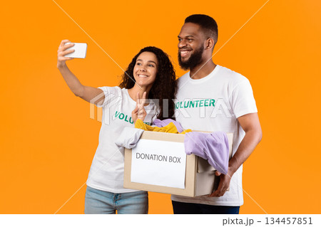 Two Multiethnic Volunteers Posing With Donation Box Making Selfie On Phone Picking Clothes For Charity Standing Over Yellow Background, Studio Shot. Contribution And Volunteering Concept Two Multiethnic Volunteers Posing With Donation Box Making Selfie On Phone Picking Clothes For Charity Standing Over Yellow Background, Studio Shot. Contribution And Volunteering Concept 134457851