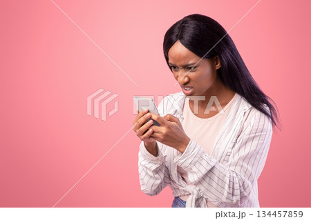 Angry black woman looking at her smartphone with disgust, reading annoying message or news on pink studio background. African American lady feeling stressed at social media publication 134457859