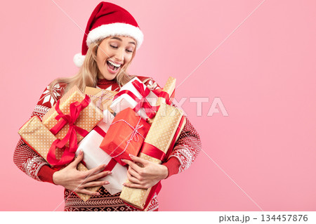 Happy Caucasian lady in cozy warm sweater and Santa hat holding heap of gifts over pink studio background. Attractive millennial woman celebrating New Year or Christmas 134457876