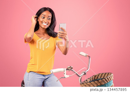 Pretty black woman on vintage bicycle taking selfie with smartphone over pink studio background. Happy African American lady on bike making photo of herself or having video chat with friend 134457895