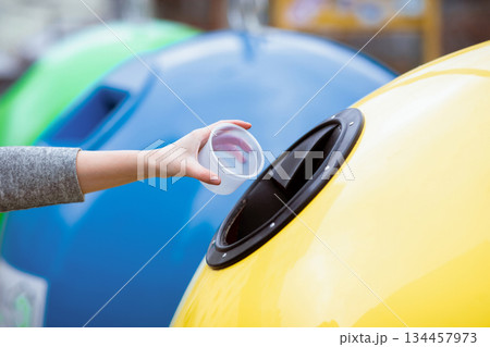 A lady hand places a plastic cup into a recycling bin in a vibrant, eco-friendly setting. The background features colorful bins promoting sustainable practices. A lady hand places a plastic cup into a recycling bin in a vibrant, eco-friendly setting. The background features colorful bins promoting sustainable practices. 134457973