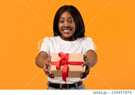 Cheerful young black lady outstretching present towards camera, orange studio background, copy space. Happy african american woman in white t-shirt giving gift box, greeting with birthday Cheerful young black lady outstretching present towards camera, orange studio background, copy space. Happy african american woman in white t-shirt giving gift box, greeting with birthday 134457974