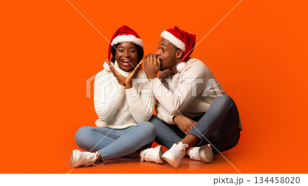 Loving Black Guy Whispering Christmas Secret To His Excited Girlfriend, Spelling To Her Ear While Having Fun Together Over Orange Background In Studio, Enjoying Celebrating Winter Holidays Together 134458020