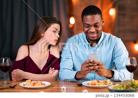 Jealous young woman peeking into boyfriend's smartphone, reading his messages during date dinner in restaurant, young black man having phubbing problem and ignoring his girlfriend, free space Jealous young woman peeking into boyfriend's smartphone, reading his messages during date dinner in restaurant, young black man having phubbing problem and ignoring his girlfriend, free space 134458066