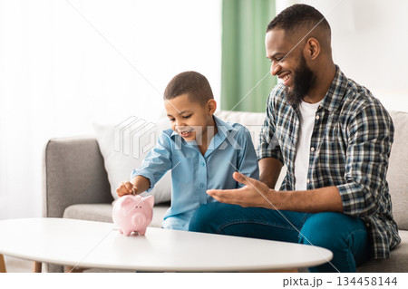 Personal Savings. African Father And Son Putting Coin In Piggybank Sitting On Sofa At Home, Dad Teaching His Kid Financial Literacy, Talking About Money And Budget Planning. Selective Focus Personal Savings. African Father And Son Putting Coin In Piggybank Sitting On Sofa At Home, Dad Teaching His Kid Financial Literacy, Talking About Money And Budget Planning. Selective Focus 134458144