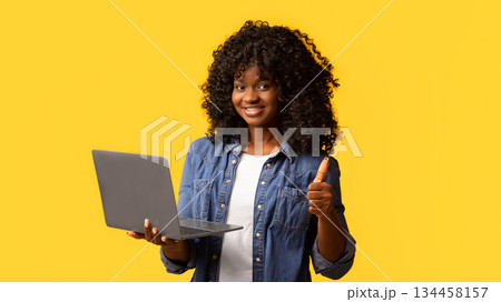 Happy african american young woman holding newest laptop and showing thumb up, yellow studio background, panorama with copy space. Cheerful black lady using notebook, shopping online or websurfing Happy african american young woman holding newest laptop and showing thumb up, yellow studio background, panorama with copy space. Cheerful black lady using notebook, shopping online or websurfing 134458157