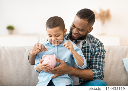 Financial Literacy. Cheerful African Father And Son Putting Money In Piggybank Sitting On Sofa At Home. Daddy Teaching His Child Budget Planning, Keeping Personal Savings Safety Financial Literacy. Cheerful African Father And Son Putting Money In Piggybank Sitting On Sofa At Home. Daddy Teaching His Child Budget Planning, Keeping Personal Savings Safety 134458169