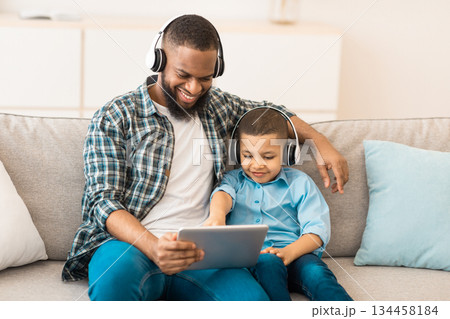 African American Dad And His Son Using Digital Tablet Wearing Headphones Watching Cartoons Online Sitting On Sofa At Home. Family Bonding Using Computer Together On Weekend African American Dad And His Son Using Digital Tablet Wearing Headphones Watching Cartoons Online Sitting On Sofa At Home. Family Bonding Using Computer Together On Weekend 134458184