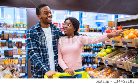 Shopping together is fun. Happy black couple choosing healthy food at fruits and veggies section of supermarket. Millennial African American family with trolley purchasing groceries at mall 134458195