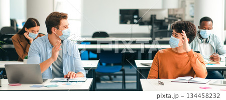 Education And Social Distancing. Smiling international male students wearing protective face masks greeting each other and waving hello at the classroom. Men sitting at desks, studying in high school Education And Social Distancing. Smiling international male students wearing protective face masks greeting each other and waving hello at the classroom. Men sitting at desks, studying in high school 134458232