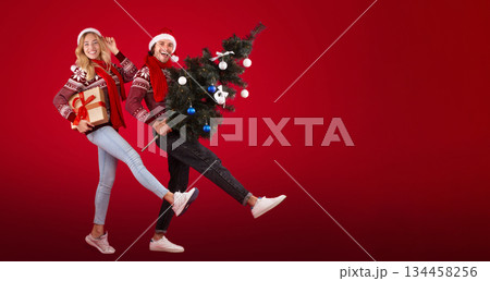 Full length portrait of young couple with Christmas gift and New Year tree walking on red background 134458256