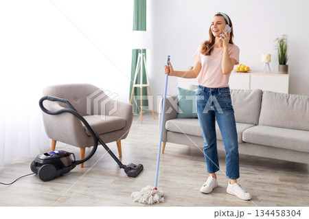 Happy young housewife speaking on smartphone during house cleanup, copy space. Cheerful millennial lady taking break from domestic duties to have phone conversation, indoors Happy young housewife speaking on smartphone during house cleanup, copy space. Cheerful millennial lady taking break from domestic duties to have phone conversation, indoors 134458304