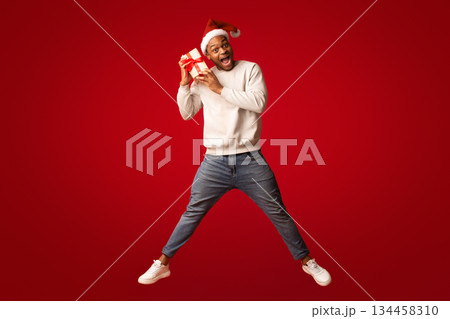 Emotional black man in santa hat jumping with christmas present box over orange background, surprised african american guy enjoying receiving xmas gifts, cheering holidays, full length, free space 134458310