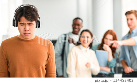 Bullying Concept. Rude diverse group of people mocking and making fun of sad asian guy wearing headphones, pointing fingers at him and laughing, standing in blurred background. Prank and abuse 134458402