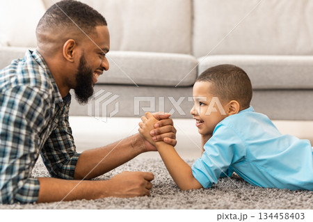 Happy African American Dad And Kid Son Arm Wrestling Competing Lying On Floor At Home. Happy Young Daddy And Little Boy Armwrestling Having Fun Together. Side View Happy African American Dad And Kid Son Arm Wrestling Competing Lying On Floor At Home. Happy Young Daddy And Little Boy Armwrestling Having Fun Together. Side View 134458403