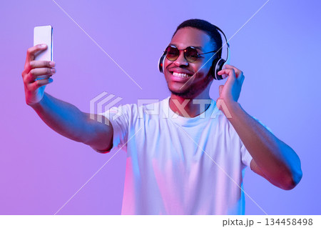 Handsome Black Man Wearing Wireless Headphones And Sunglasses Taking Selfie On Smartphone, Cheerful African American Guy Posing At Camera While Standing In Neon Lighting Over Purple Background 134458498