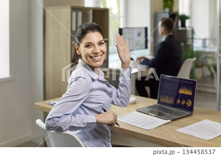 Happy office employee waving at camera near laptop with analytics charts on desk 134458537