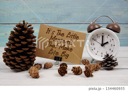 It's The Little Moments That Make Life Big text written on paper card with alarm clock and pine cones decoration on wooden background 134460935