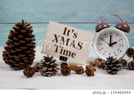 It's XMAS Time text written on paper card with alarm clock and pine cones decoration on wooden background It's XMAS Time text written on paper card with alarm clock and pine cones decoration on wooden background 134460944