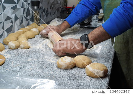 Making unleavened flatbreads. A man's hands roll out roti dough with a rolling pin. The surface is dusted with multigrain flour. Several balls of dough lie nearby. A gas stove is in the background. Making unleavened flatbreads. A man's hands roll out roti dough with a rolling pin. The surface is dusted with multigrain flour. Several balls of dough lie nearby. A gas stove is in the background. 134462347