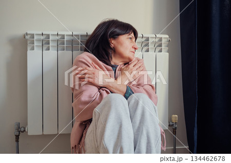 Woman shivering from cold temperatures embracing herself, sitting by radiator in her home Woman shivering from cold temperatures embracing herself, sitting by radiator in her home 134462678