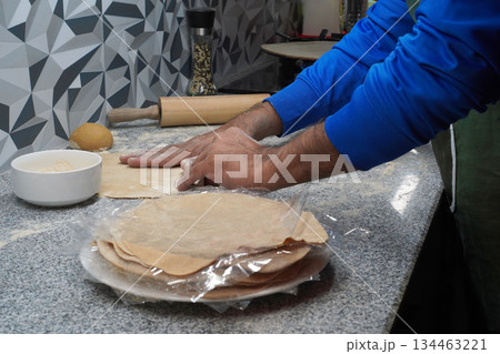 Making unleavened flatbreads. A man's hands touch a flatbread. Multigrain flour, a ball of dough and a rolling pin lie nearby. A stack of flatbreads is ready to freeze. A gas stove with bread baking. Making unleavened flatbreads. A man's hands touch a flatbread. Multigrain flour, a ball of dough and a rolling pin lie nearby. A stack of flatbreads is ready to freeze. A gas stove with bread baking. 134463221
