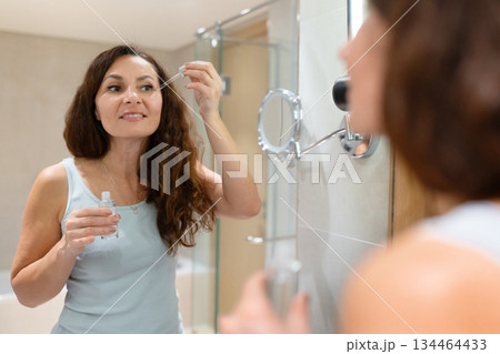 Adult caucasian woman applying cosmetic serum under eyes near bathroom mirror 134464433